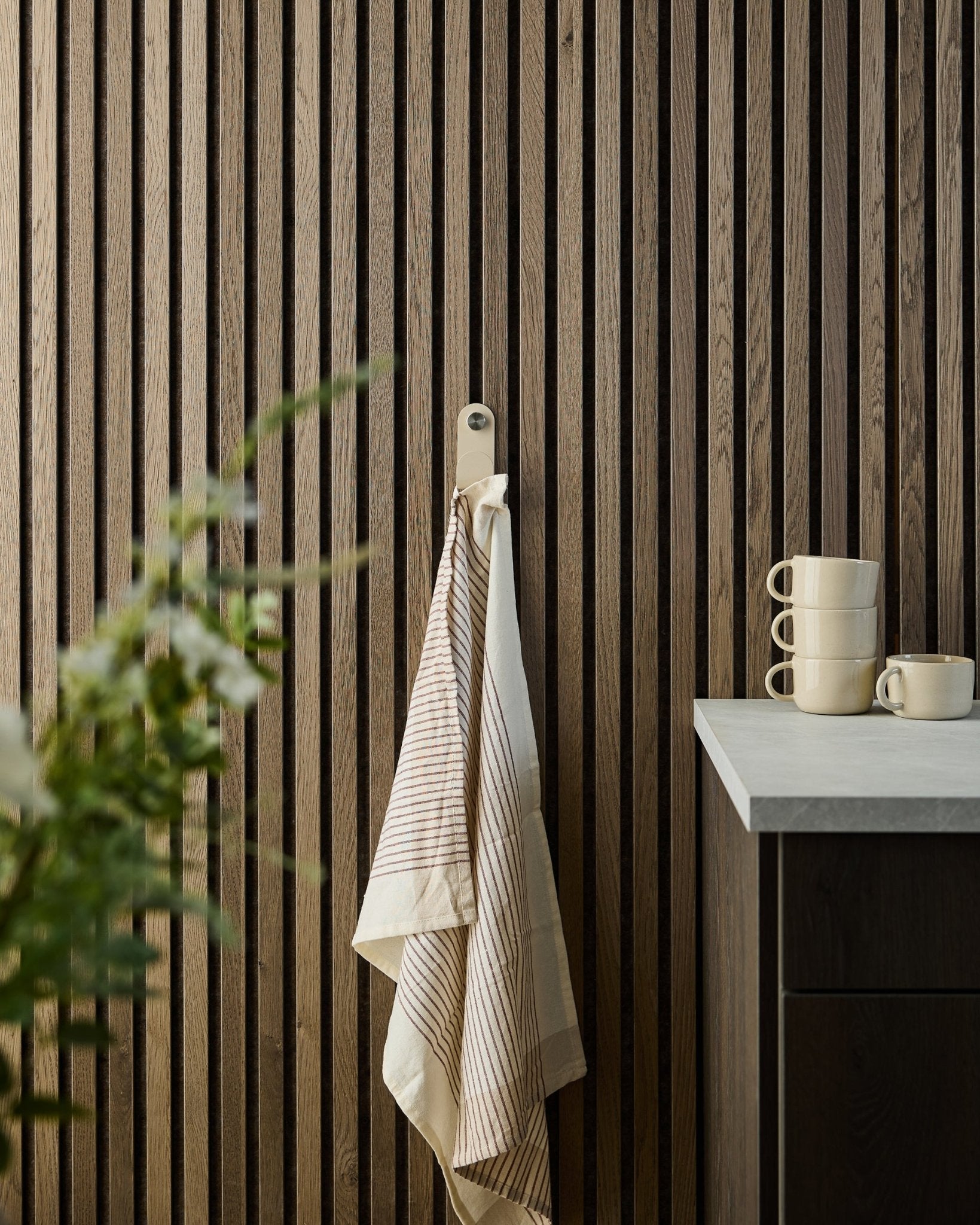 En randig handduk hänger på en beige WoodUpp Norr Single Hanger monterad på en trälamellpanel Akustikpanel; staplade muggar och en växt står på bänken nedanför.
